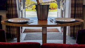 A dining table with two plates and cutlery, a bottle of champagne, and a view of a vineyard through open doors. - Field House Bothy - UK12571 (Borrowdale, near Keswick)