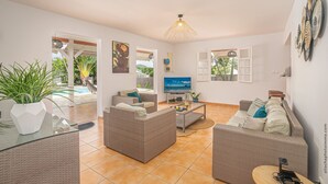 Amplia sala de estar con mobiliario de mimbre, un televisor de pantalla plana y un ventanal con vistas a una piscina.