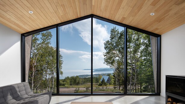A modern living room with a large glass wall offering a scenic view of trees and mountains. - Le Littoral : Luxurious Chalet with Pool Sauna Spa & View (La Malbaie)