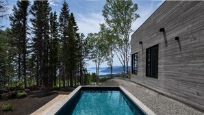 A modern building with a wooden exterior and a rectangular pool in the foreground. - Le Littoral : Luxurious Chalet with Pool Sauna Spa & View (La Malbaie)