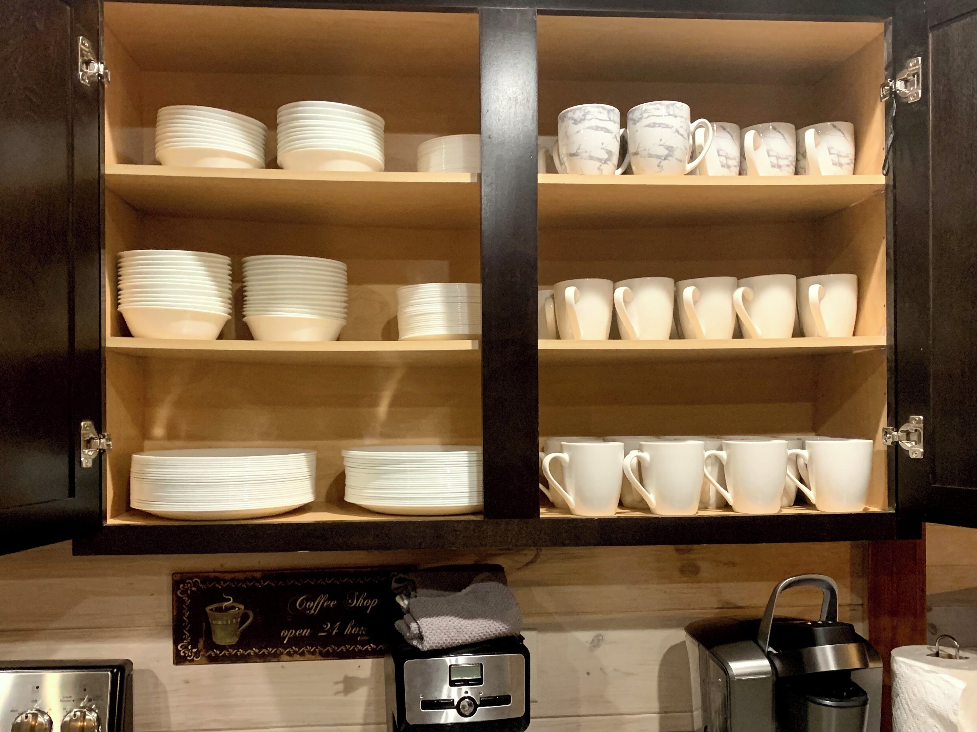 A kitchen cabinet with shelves holding white plates and cups. There is a coffee shop sign on the wall and a kettle on the counter.