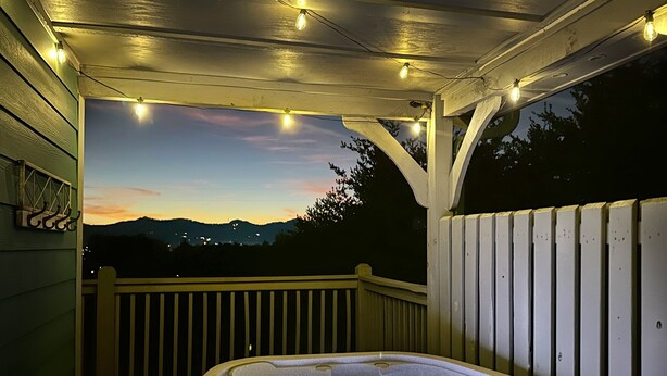 A hot tub on a wooden deck with string lights overhead, overlooking a mountainous landscape.