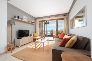 A modern living room with a flat-screen TV, a sofa, a coffee table, and a balcony with a view of the sea.
