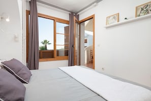 A bedroom with a bed, a window with curtains, a small shelf with pictures, and a view of the outside.