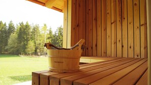 Ein aus Holz gefertigter Eimer mit dem Aufdruck 'WASSERFELD' auf einer Holzbank, mit Blick auf ein grĂŒnes Feld und BĂ€ume im Hintergrund.
