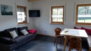 A living room with a dark blue sofa, a wooden dining table, and a TV mounted on the wall. - Ferienwohnung Schmalzgrub (Böbrach)