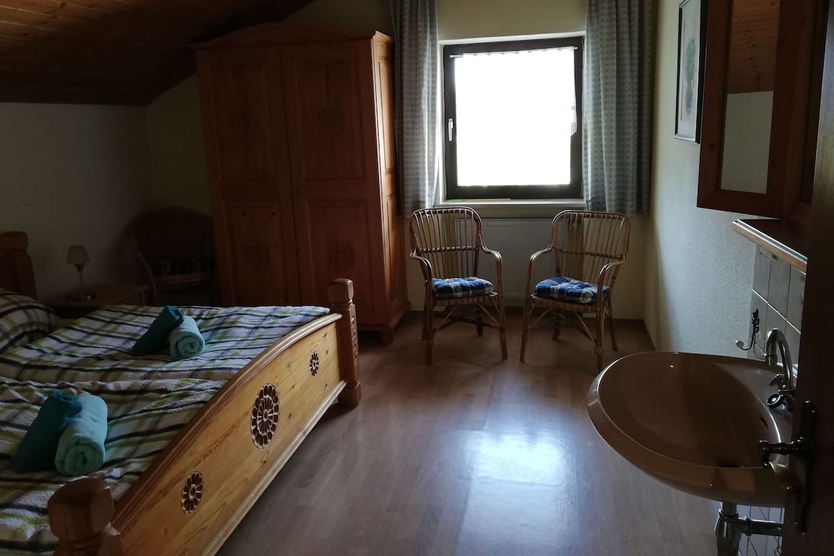 A bedroom with a wooden bed, a wooden wardrobe, two wicker chairs, a window with curtains, and a bathroom with a sink.