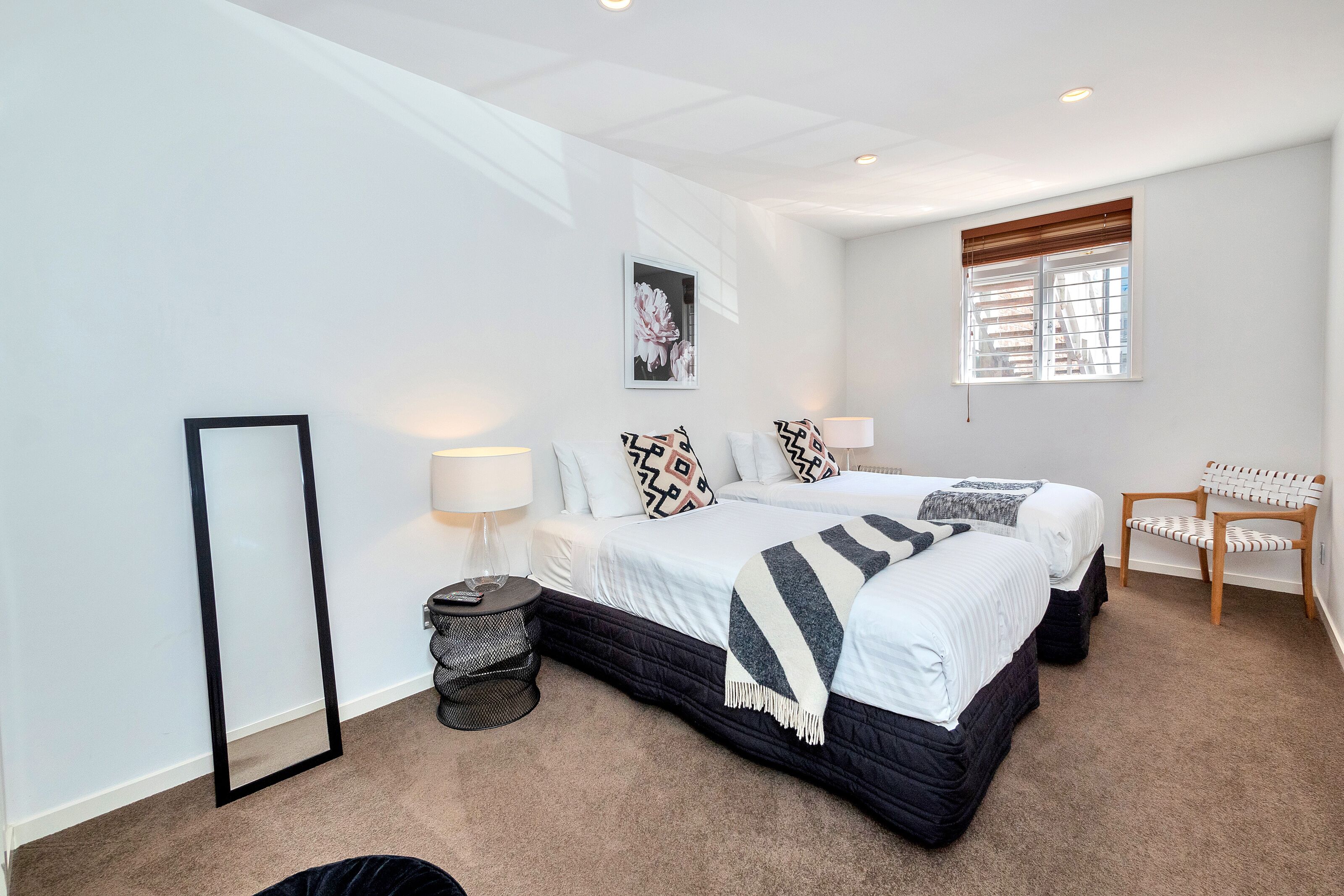 Een slaapkamer met twee bedden, een raam met zonwering, een stoel, een lamp en een lijst met een afbeelding aan de muur.