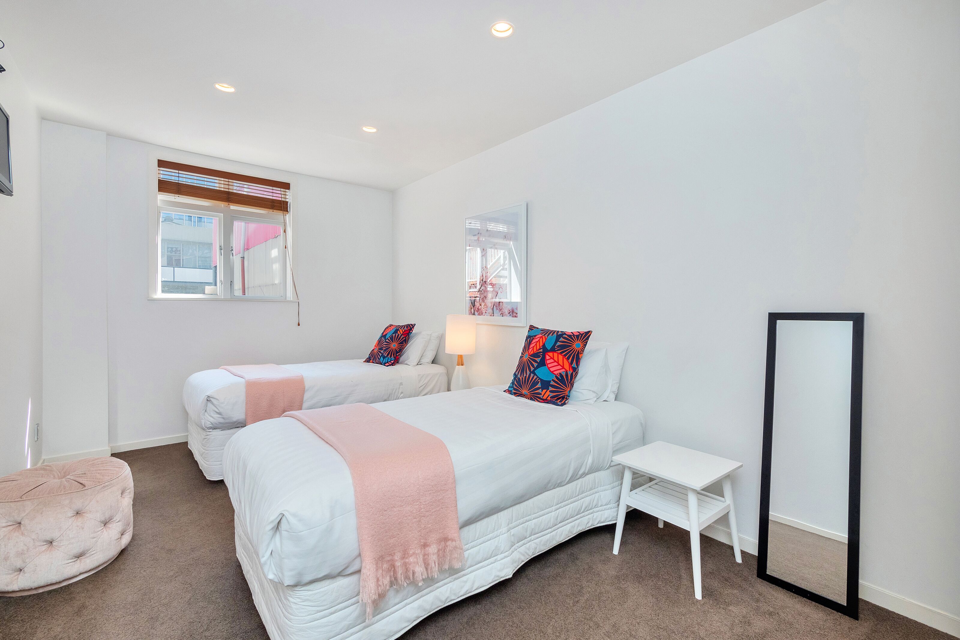 A bedroom with two beds, a small table, a mirror, and a window with blinds.