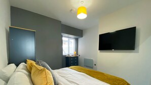 A modern bedroom with a bed, a bedside table, a window, and a flat-screen TV mounted on the wall. - Chelsea Suite Sasco Apartments (Blackpool)