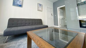 A modern living room with a grey sofa, a glass-top coffee table, and a kitchen area with a grey cabinet and white door. - Chelsea Suite Sasco Apartments (Blackpool)