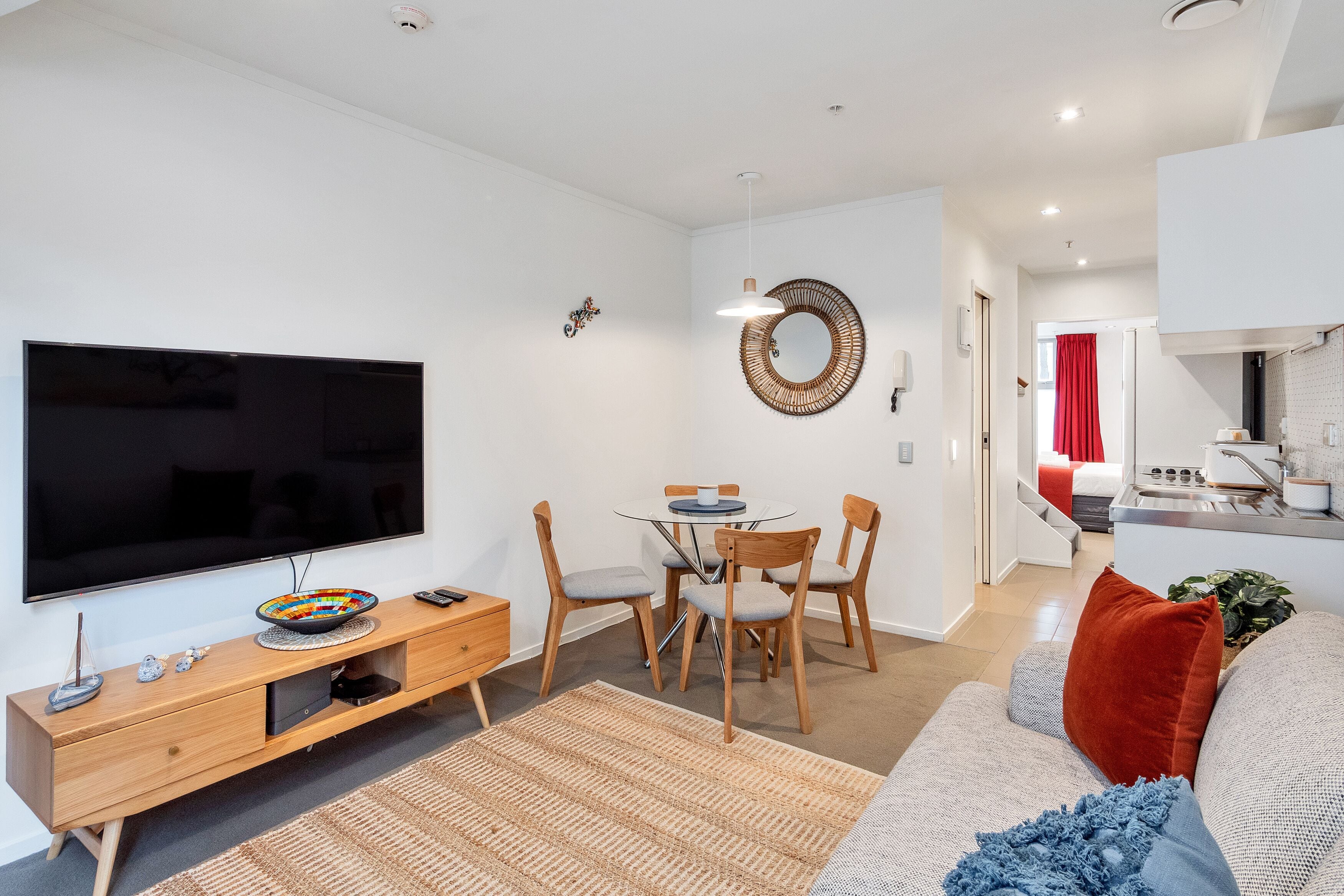 A modern living room with a flat-screen TV, wooden furniture, a dining table, and a kitchen area with red curtains.