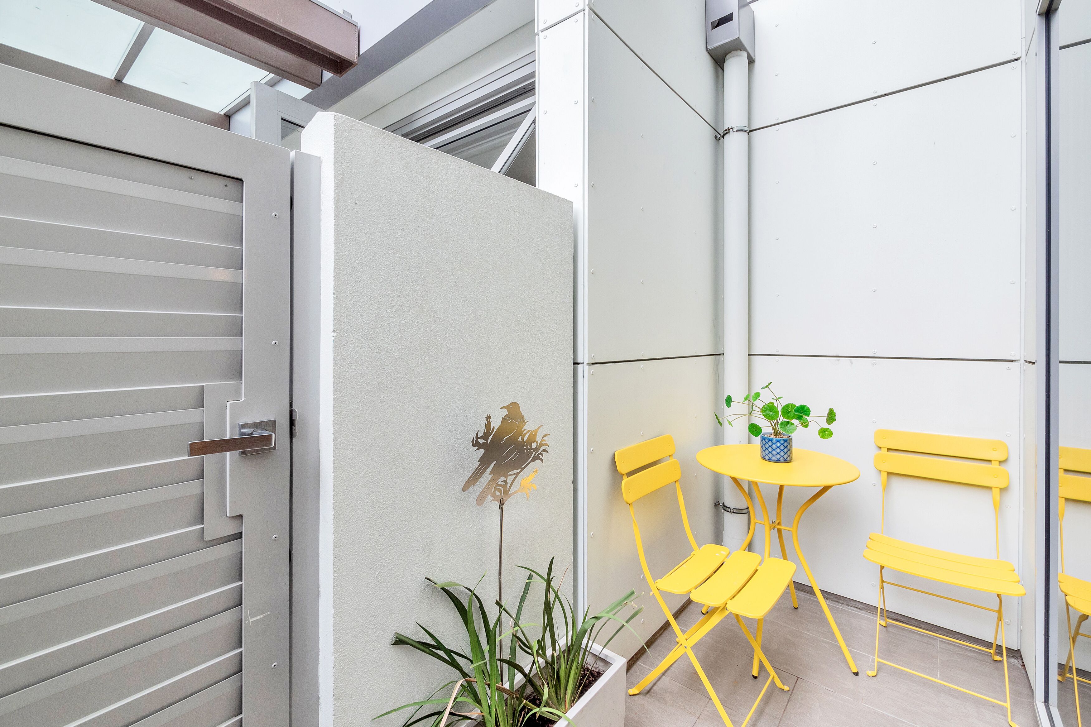 Un área de estar moderna al aire libre con sillas amarillas y una mesita, una pieza decorativa de metal en la pared y una planta en maceta.
