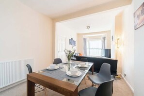 A dining area with a glass table, four chairs, a vase with flowers, and a sofa in the background.