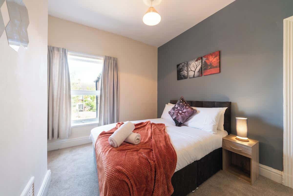 A hotel room with a bed, a nightstand, a lamp, a window with curtains, and a wall art piece.