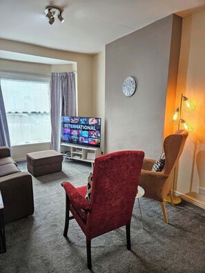 A living room with a television displaying 'INTERNATIONAL', a red armchair, a brown sofa, a small ottoman, and a lamp.