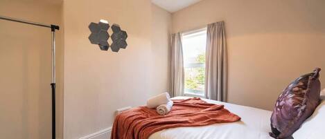 A hotel room with a bed, a window with curtains, a wall-mounted artwork, and a ceiling light.