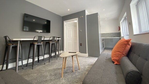 A modern living room with a grey sofa, a TV mounted on the wall, and a dining area with a table and chairs. - Kensington Suite Sasco Apartments (Blackpool)