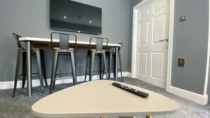 A room with a flat-screen TV mounted on the wall, a beige table with wooden legs, and four black bar stools with wooden accents. - Kensington Suite Sasco Apartments (Blackpool)