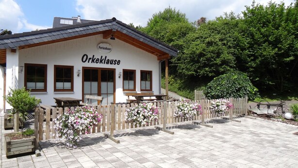 Een gebouw met de naam "Orkeklaus", omringd door een houten hek, bloembedden en een verharde oprijlaan.
