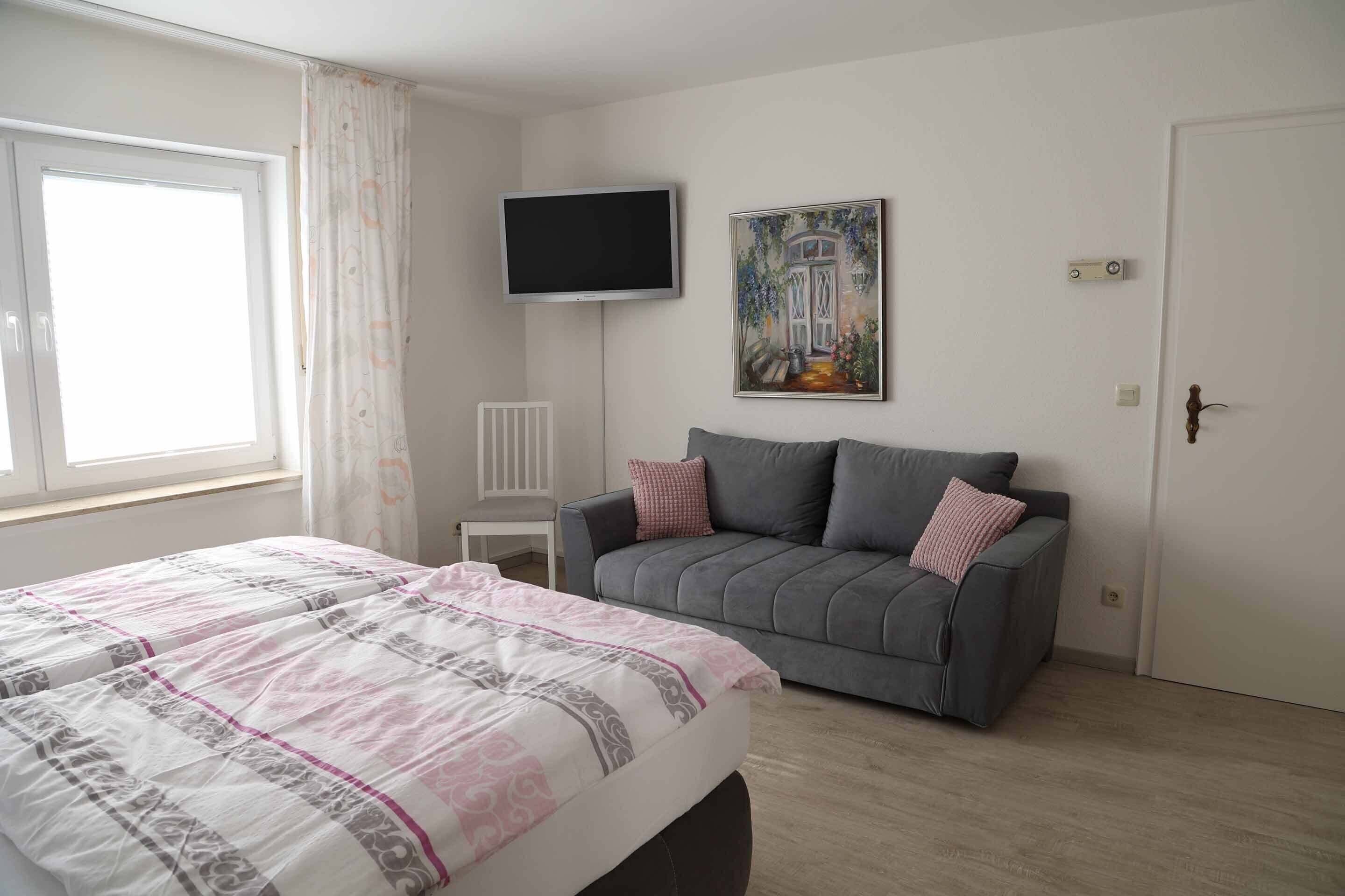 A bedroom with a bed, a grey sofa, a TV, and a painting on the wall.