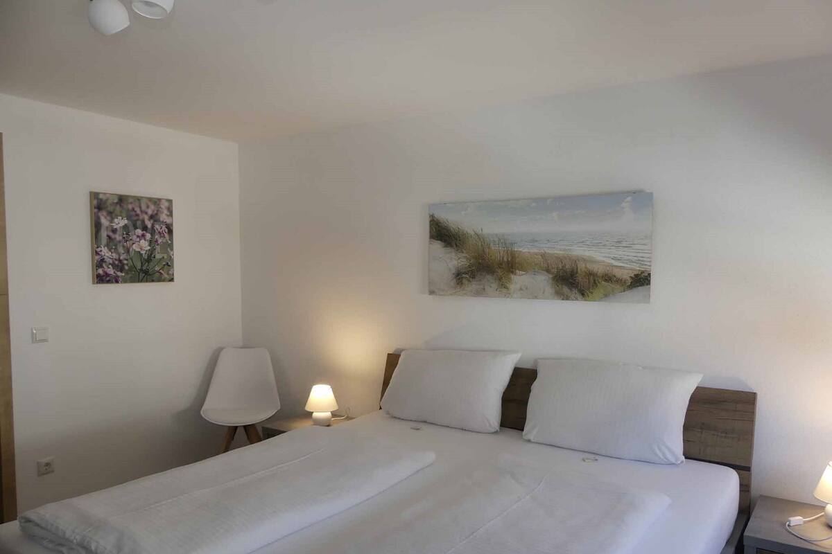 A neatly made bed with white linens, a bedside lamp, and a painting of a beach scene on the wall.