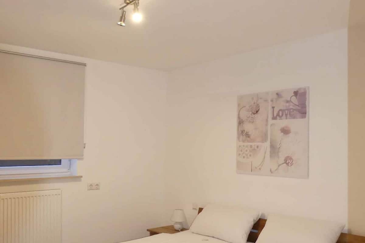 A bedroom with a double bed, a nightstand, a wall-mounted light, and a window with blinds.