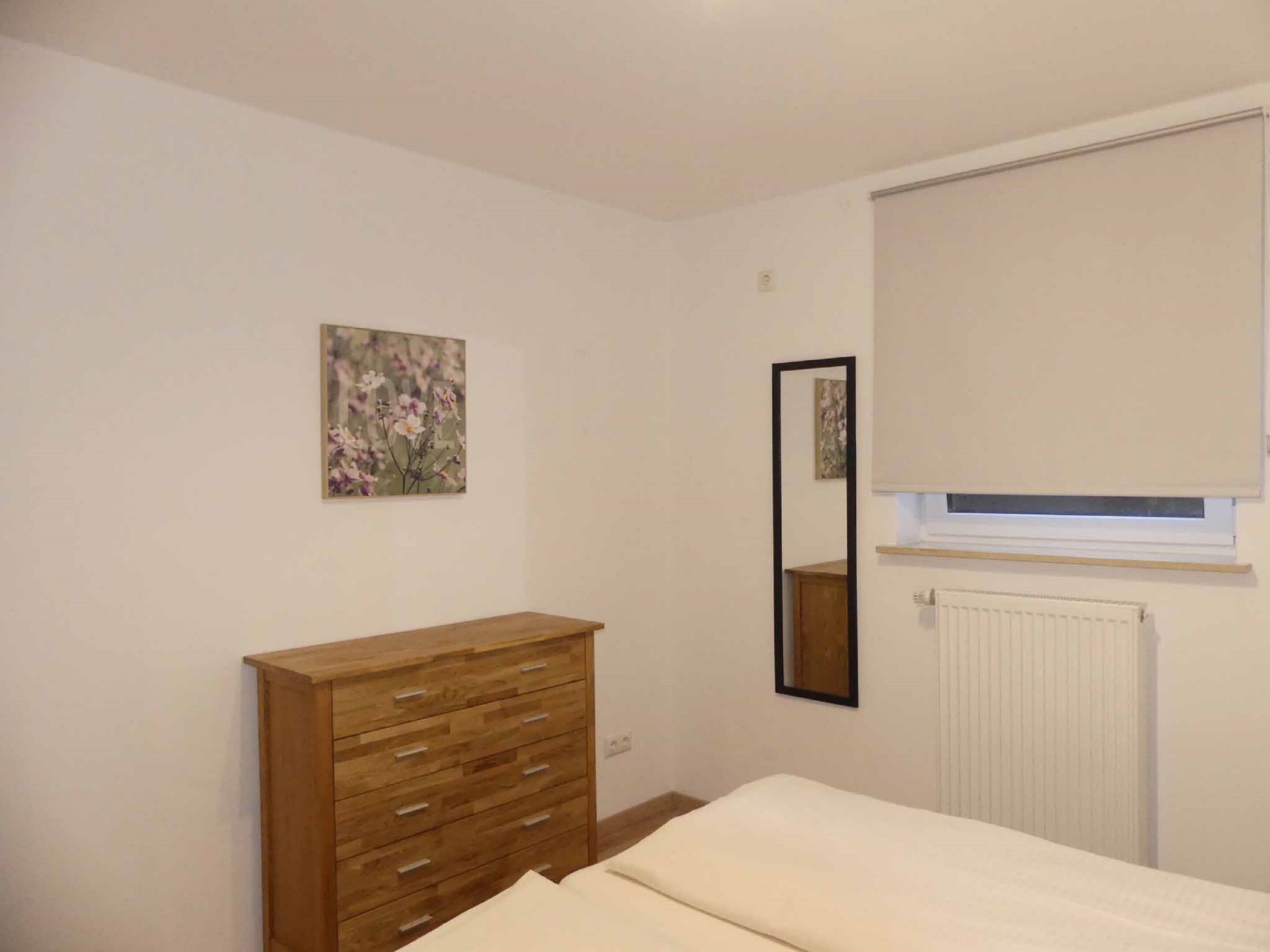 A bedroom with a wooden dresser, a bed, a window with a blind, a radiator, and a wall-mounted mirror.