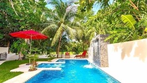 A swimming pool with a waterfall feature, surrounded by lush greenery and palm trees. - Villa Corazon Deluxe (Puerto Aventuras)
