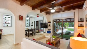 A spacious living room with a sofa, a coffee table, and a TV. There is a dining area with chairs and a table, and a sliding glass door leading to a patio. - Villa Corazon Deluxe (Puerto Aventuras)