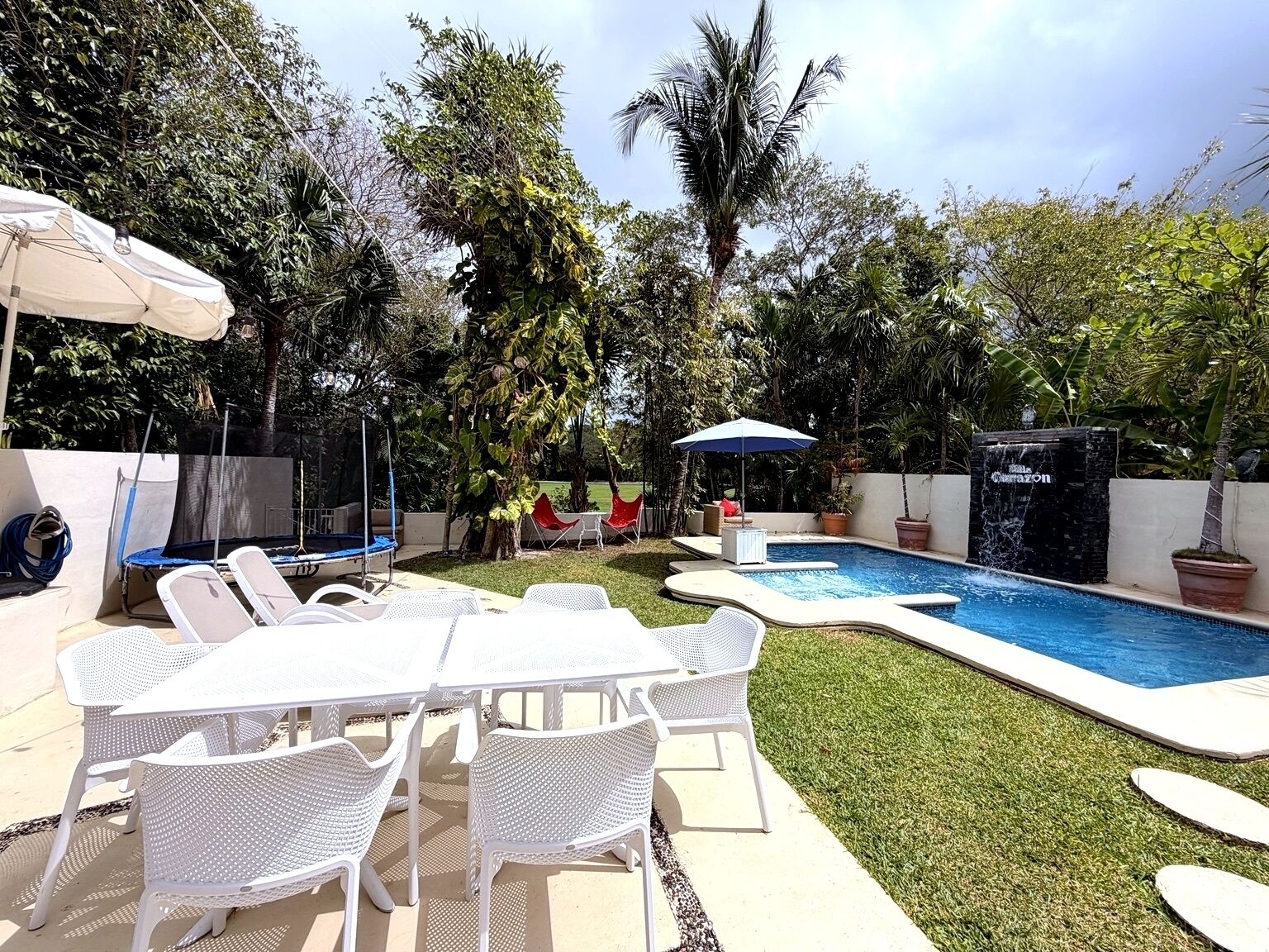Um quintal com piscina, móveis de jardim brancos e vegetação exuberante.