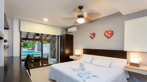 A bedroom with a bed, bedside tables, a ceiling fan, and a view of an outdoor pool. - Villa Corazon Deluxe (Puerto Aventuras)