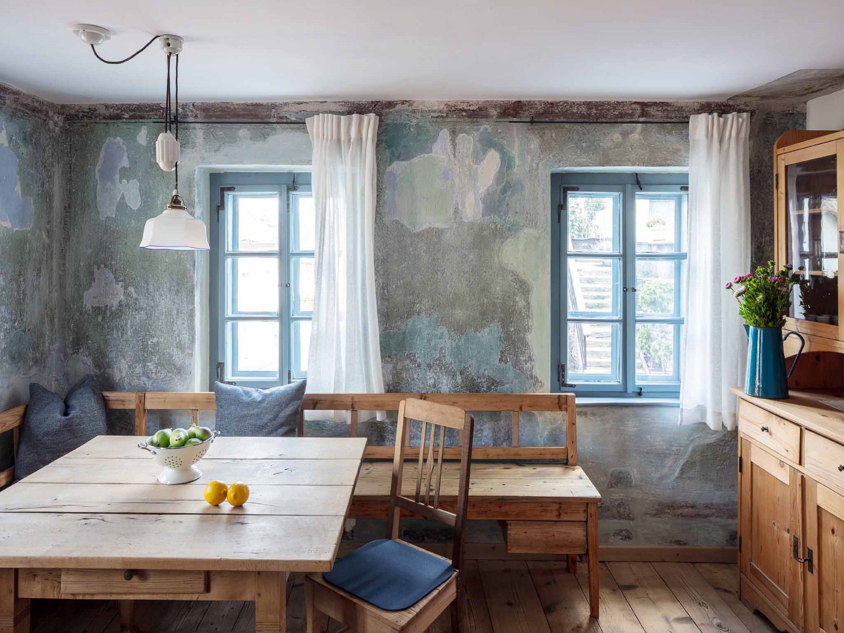 A rustic kitchen with wooden furniture, a table with fruits, and two windows with curtains.