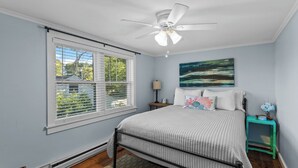 A bedroom with a bed, a ceiling fan, a window with blinds, a nightstand, and a painting on the wall.