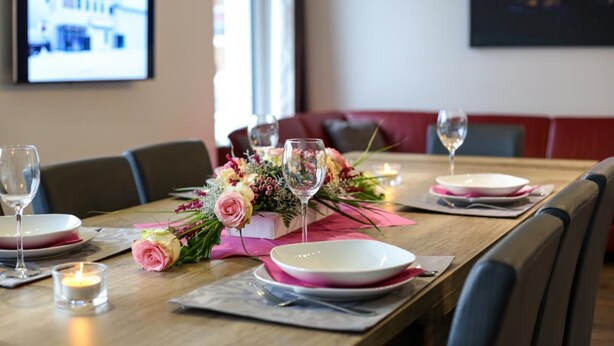 Ein Esstisch ist gedeckt mit Tellern, Gläsern und einer Blumenarrangememt.