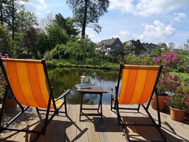 Duas cadeiras laranja viradas para um deck de madeira com uma mesa e um pequeno lago.