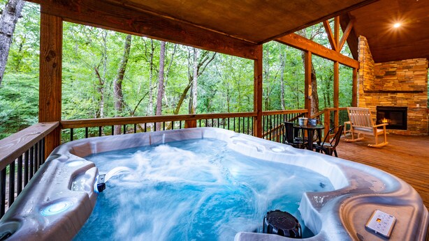 A hot tub on a wooden deck with a stone fireplace and a view of a forest. - Up Chic Creek: Sleeps 6, Creekside, Swim Hole, Fireplaces, Hot Tub, Pet Friendly (Broken Bow)