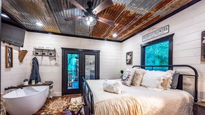 A rustic bedroom with a large white bathtub, a bed with white linens, a ceiling fan, and a window with a view of trees. - Up Chic Creek: Sleeps 6, Creekside, Swim Hole, Fireplaces, Hot Tub, Pet Friendly (Broken Bow)