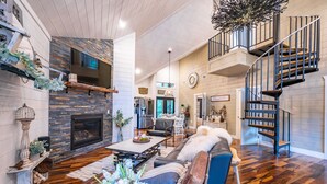 A spacious living room with a fireplace, a staircase, and a kitchen in the background. - Up Chic Creek: Sleeps 6, Creekside, Swim Hole, Fireplaces, Hot Tub, Pet Friendly (Broken Bow)