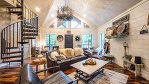 A spacious living area with a black leather sofa, a coffee table, and a wooden staircase. - Up Chic Creek: Sleeps 6, Creekside, Swim Hole, Fireplaces, Hot Tub, Pet Friendly (Broken Bow)