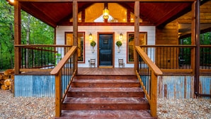 A wooden staircase leading to a porch with a door, two wall-mounted lights, and a hanging light fixture. - Up Chic Creek: Sleeps 6, Creekside, Swim Hole, Fireplaces, Hot Tub, Pet Friendly (Broken Bow)