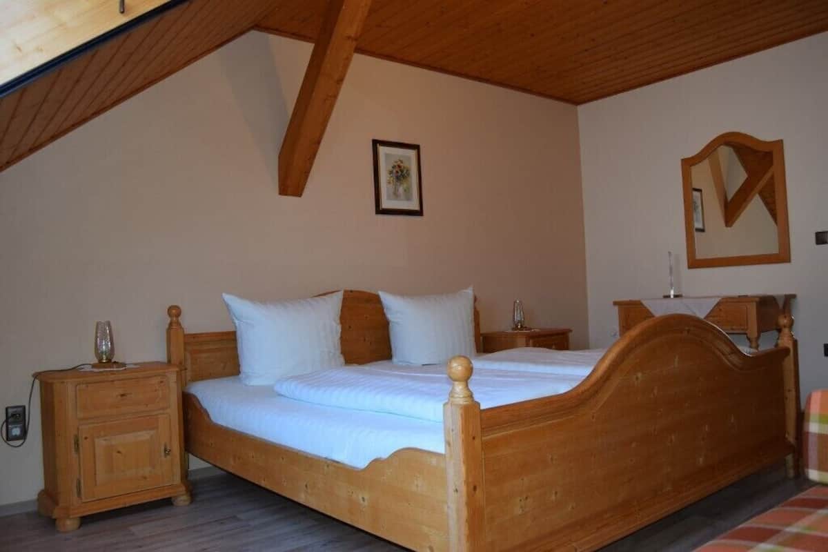 A wooden bed in a room with a sloped ceiling, a nightstand, a bedside table, and a mirror on the wall.