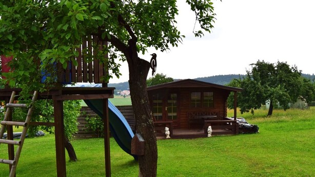 A wooden cabin with a porch and a slide in a grassy area. - Ferienhaus Herzog (Haundorf)