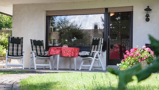 Un patio con dos sillas blancas, una mesa con mantel rojo y un vidrio que da a un edificio.