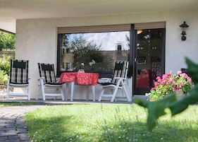 Un patio con dos sillas blancas, una mesa con mantel rojo y un vidrio que da a un edificio.