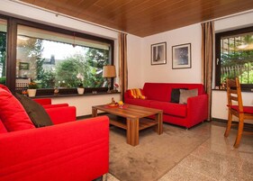 Una sala de estar moderna con muebles rojos, una mesa de centro de madera y ventanas amplias con cortinas.