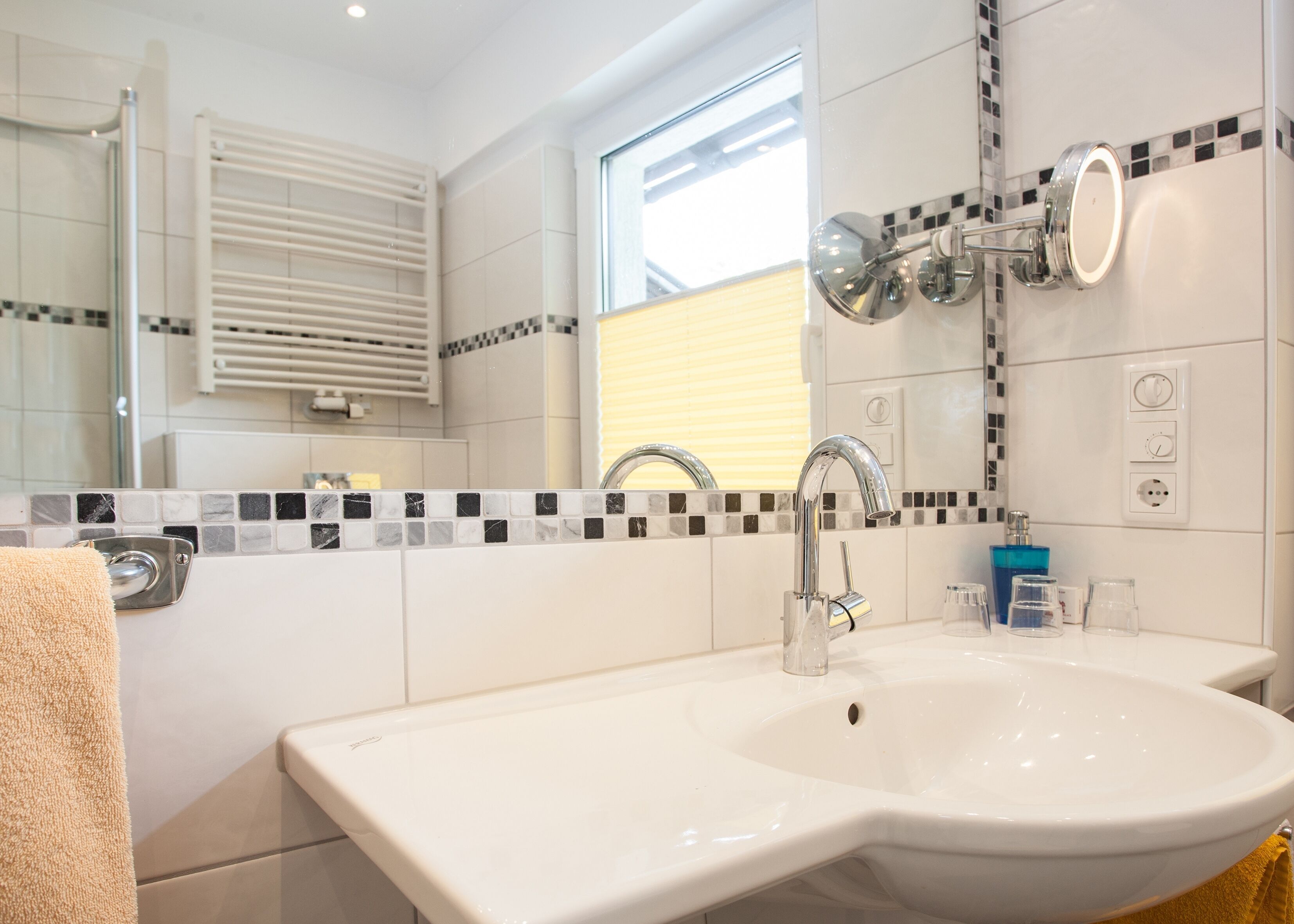 Een badkamer met een wit wastafel, een chromen kraan en een spiegel. Er is een raam met zonwering, een handdoekhouder en een betegelde spatwand.