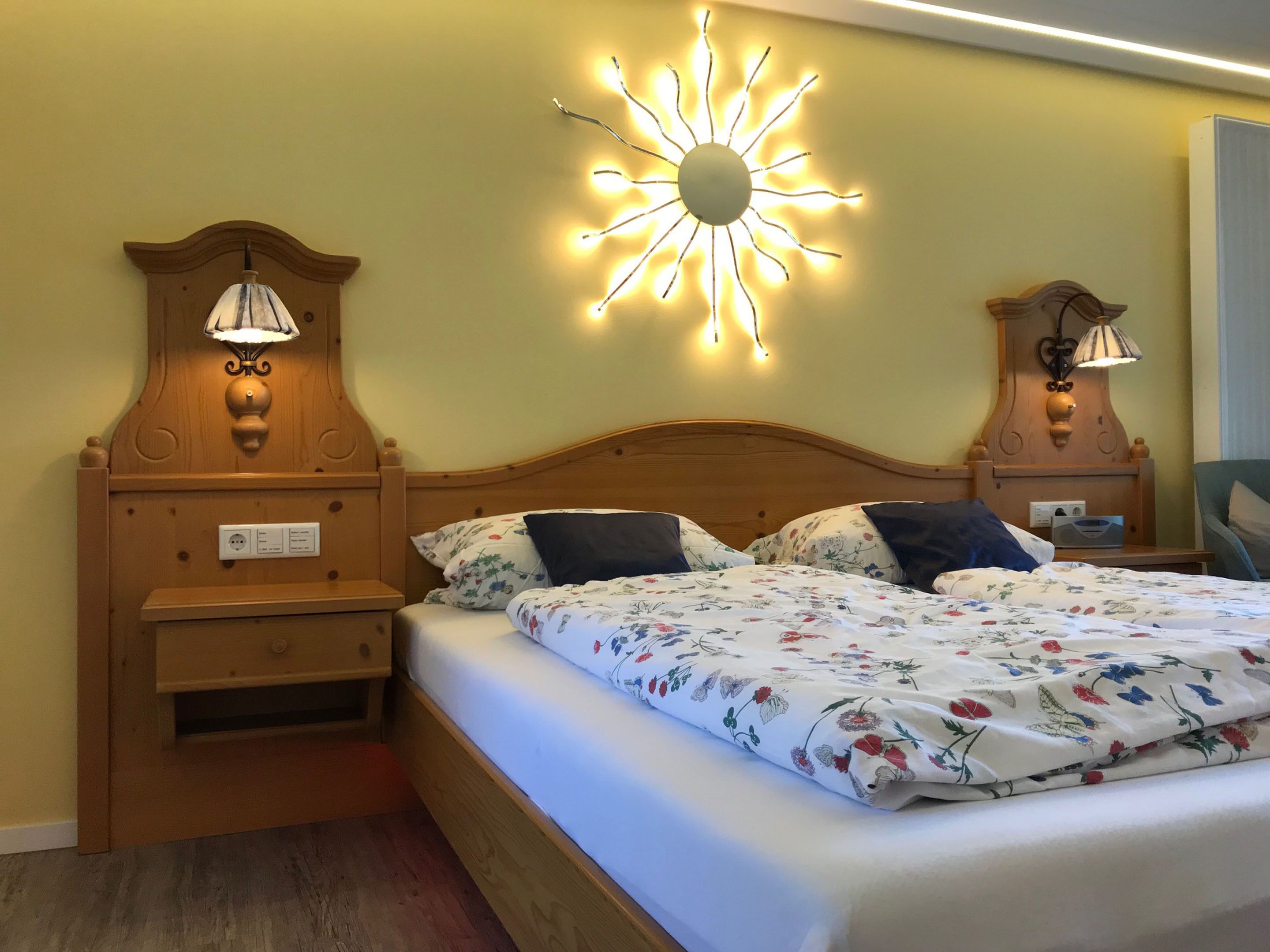 Una cama bien hecha con ropa de cama floral, dos mesitas de noche con lámparas y un accesorio de iluminación empotrado en la pared con forma de sol.