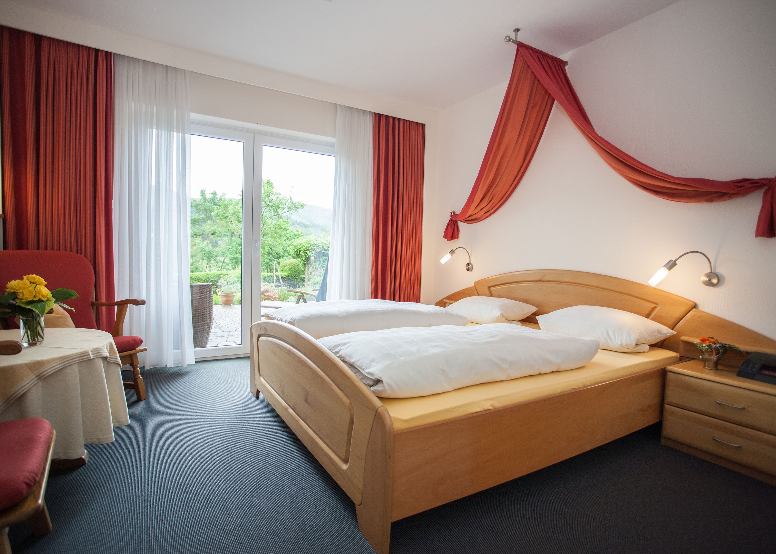 Ein Hotelzimmer mit Bett, Schreibtisch und Stuhl. Ein Fenster mit Vorhängen, eine Lampe und Blick ins Grüne sind ebenfalls vorhanden.
