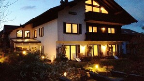 Una casa de dos pisos con un amplio porche delantero, varios ventanales e iluminación exterior.